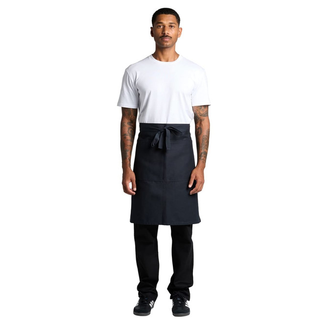 A man in a white t-shirt wears a dark grey Carrie Half Apron, with a tied waist and a logo on the apron.
