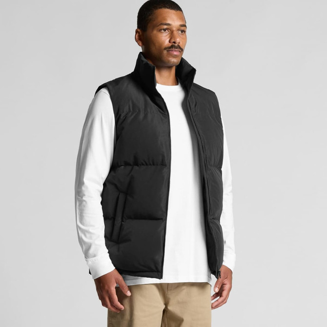 A man in a black puffer vest over a white long-sleeve shirt, standing against a neutral background.