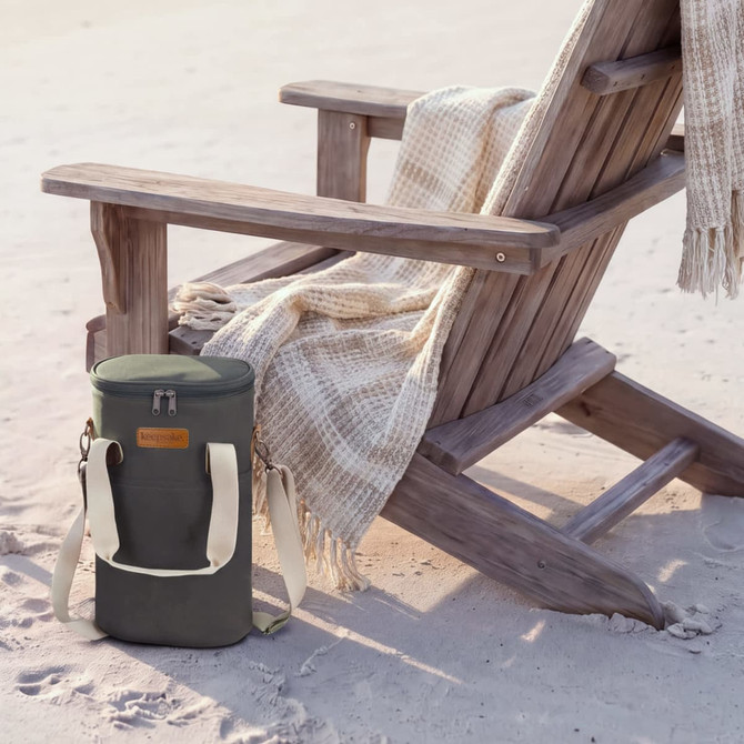 A wine cooler bag in olive green with a shoulder strap rests on the sand beside a wooden chair and a blanket.