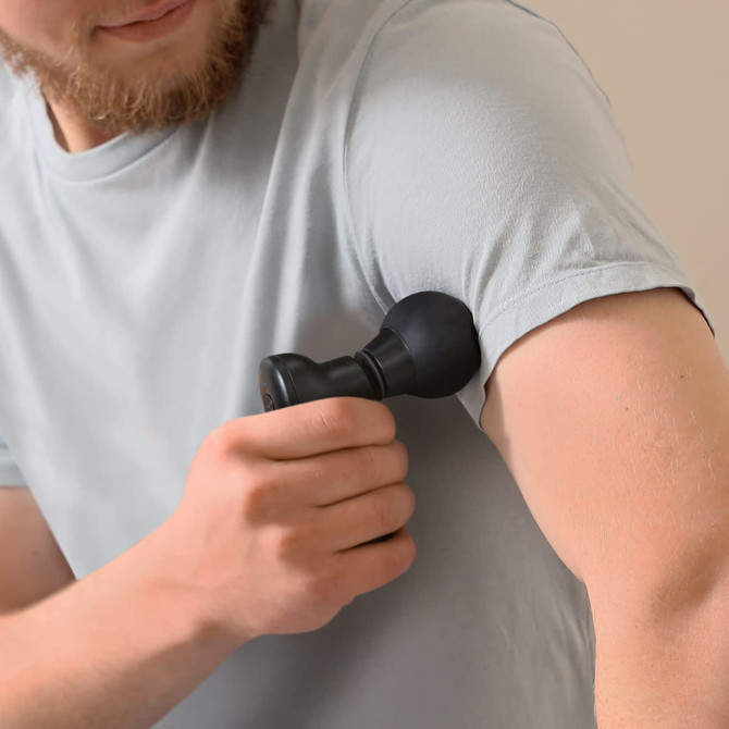 A compact handheld massager in black is being used on a person's arm, with a light-coloured t-shirt visible.