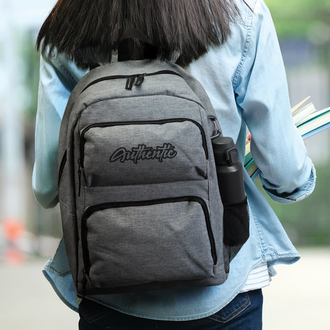 A grey backpack with a side mesh pocket for a drink bottle, carried by a person holding books.