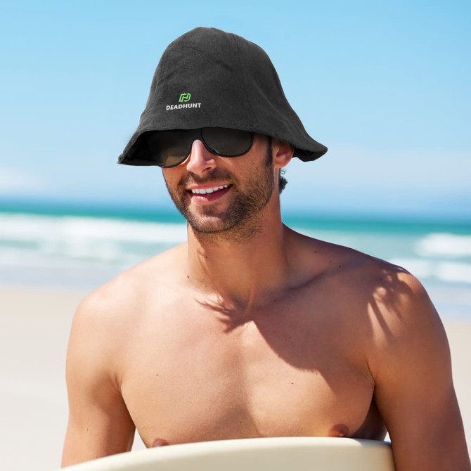 A man wearing a black bucket hat and sunglasses, sitting on a beach with ocean waves in the background.