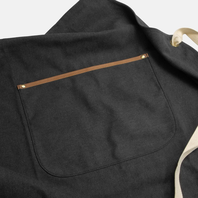 A dark grey canvas apron featuring a back pocket with a tan leather trim and an adjustable strap.