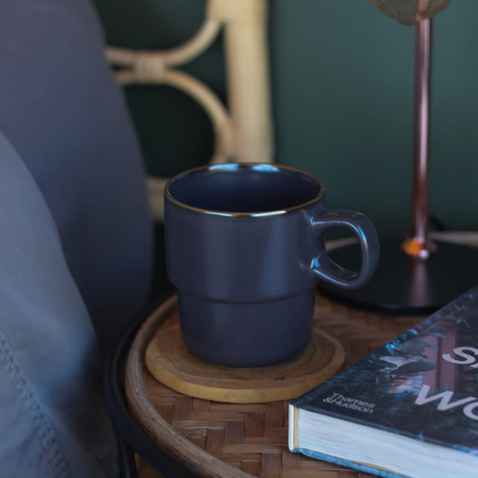 A grey stackable coffee cup sits on a wooden coaster next to a book and a lamp. The cup has a gold rim and a handle.