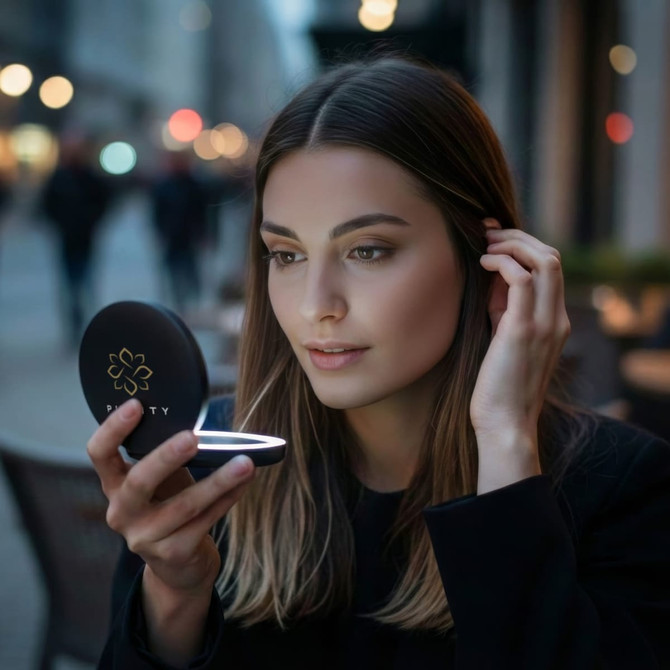 A compact mirror with LED lights, featuring a black exterior. A woman is using it in a softly lit urban setting.