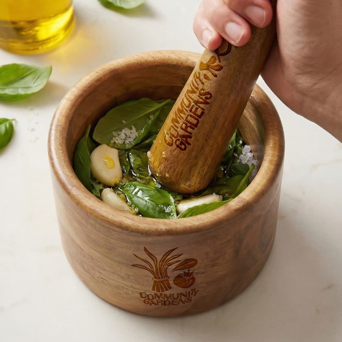 A wooden mortar and pestle filled with fresh basil, garlic, salt, and oil, featuring a logo on the side.