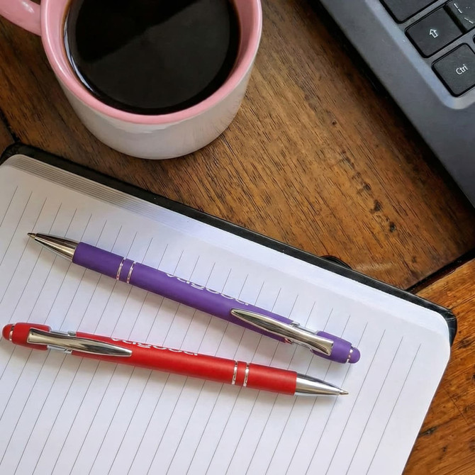 A red and a purple stylus pen rest on a white lined notepad beside a pink coffee mug and a computer keyboard.
