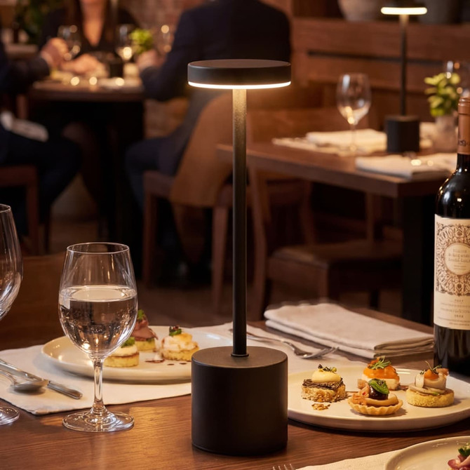A black rechargeable table lamp stands on a dining table, illuminating an elegant setting with food and drinks.