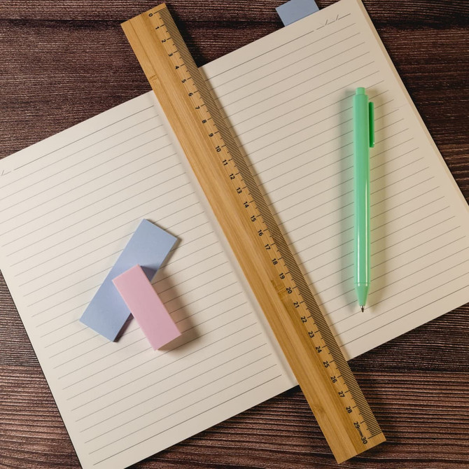 A bamboo 30cm ruler lies across a notebook, accompanied by a green pen and pink and blue sticky notes.