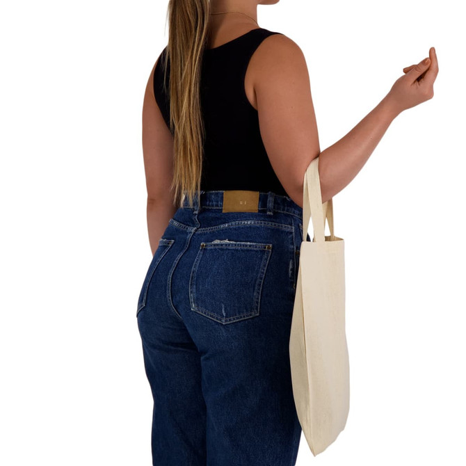 A person carrying a natural-coloured tote bag with short handles while wearing a black top and denim jeans.