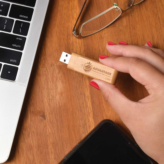 A bamboo USB flash drive held in a hand, resting on a wooden surface beside a laptop and glasses.