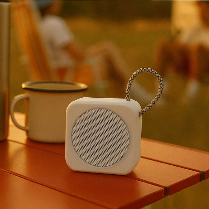 A compact white speaker with a woven strap, placed on a wooden table near a drink bottle.