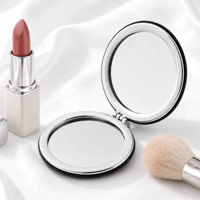 A compact mirror with a sleek silver frame, accompanied by a lipstick and a makeup brush on a soft white fabric background.