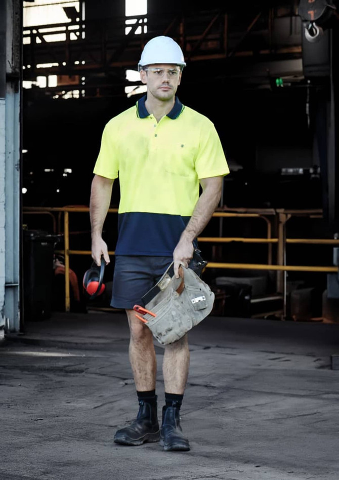 A man wearing a high-vis yellow and navy polo shirt, shorts, and a hard hat, holding work gear in a warehouse setting.
