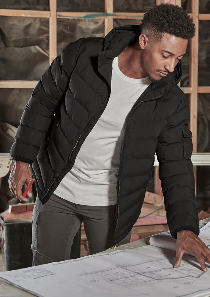 A black unisex puffer jacket with a hood, worn by a person examining blueprints indoors. The jacket has a logo.