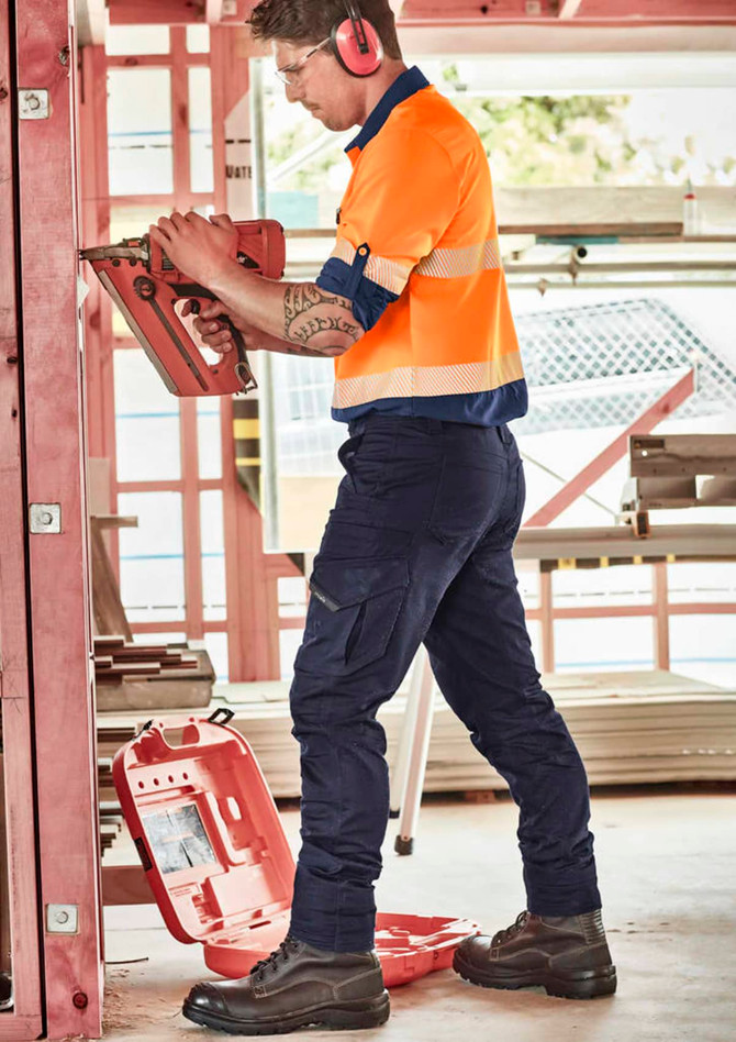 A worker in high-visibility orange shirt and navy cargo pants uses a tool on a construction site. Features branding.