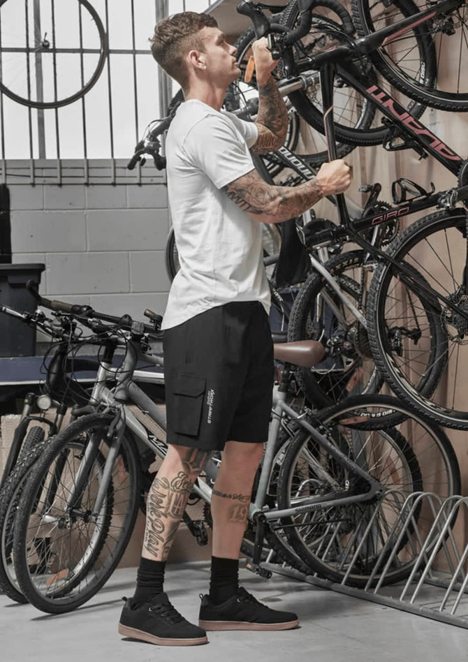 A man stands in a garage adjusting a bike, wearing black Men's Streetworx board shorts and a white t-shirt.