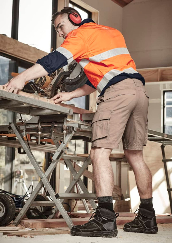 A man wearing beige cargo shorts and a high-visibility orange shirt works with power tools in a construction setting.