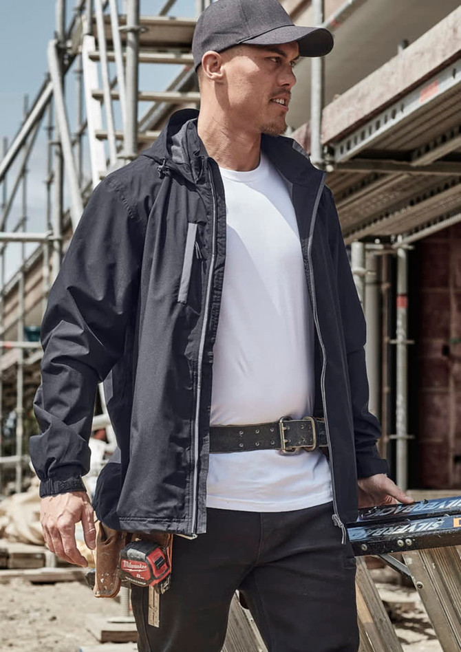 A man in a black packable rain jacket stands at a construction site, wearing a cap and tool belt.