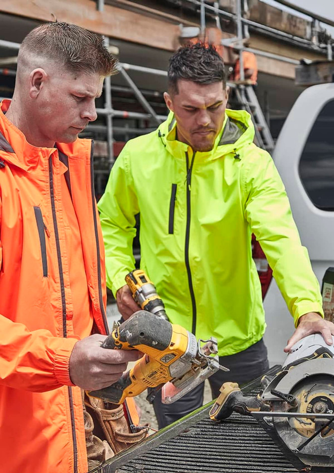 Two men in bright orange and neon yellow rain jackets work on a construction site, using power tools. Both jackets have logos.