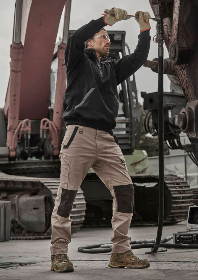 A man wears a black pullover while working with machinery, paired with beige and brown pants, in an industrial setting.