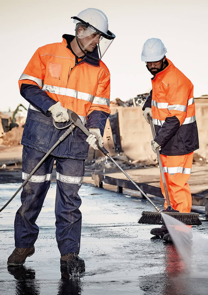 Two men in orange high-visibility jackets and helmets, working with tools on a construction site.