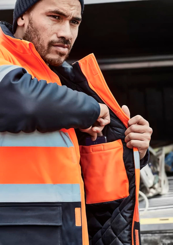 A man in an orange and navy softshell jacket with reflective strips, reaching into an inner pocket.