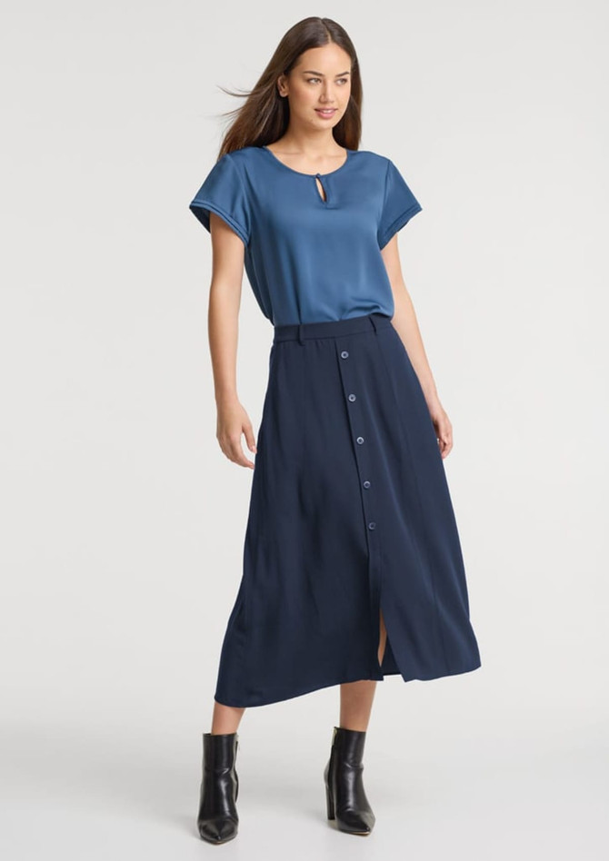A woman wearing a blue top and a navy skirt with buttons, paired with black ankle boots. The skirt features a front split.