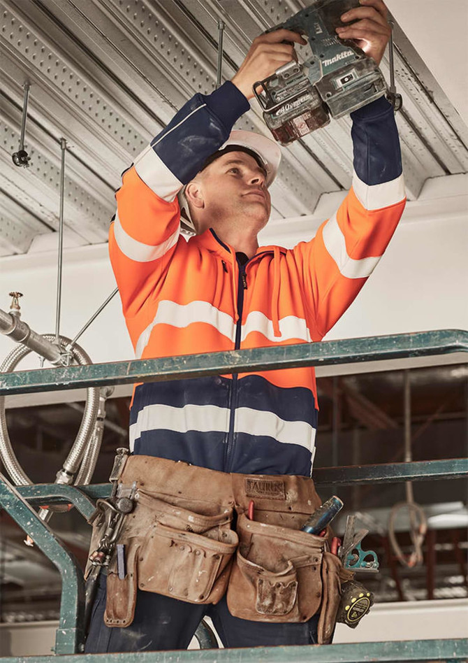 A worker in an orange and navy hi-vis full zip hoodie uses a tool while standing on a scaffold, wearing a hard hat.