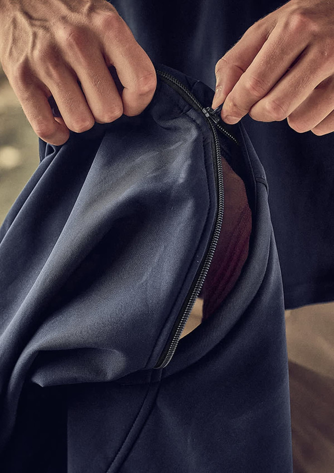 A close-up of hands zipping a navy softshell jacket with a contrasting inner layer. The jacket features a logo.