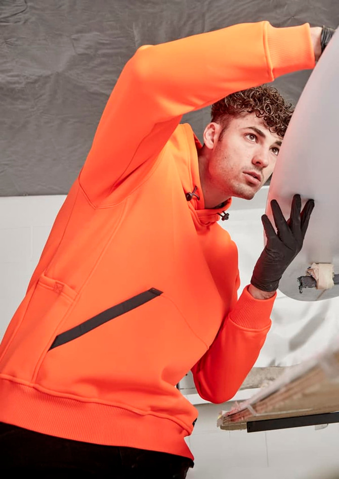 An orange unisex water-resistant hoodie featuring a front pocket and a logo, worn by a person working on a surfboard.