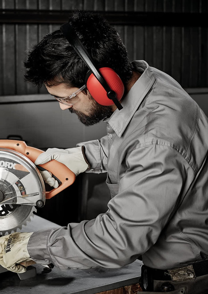 A man in a grey long sleeve work shirt uses a power saw, wearing protective earmuffs and gloves.