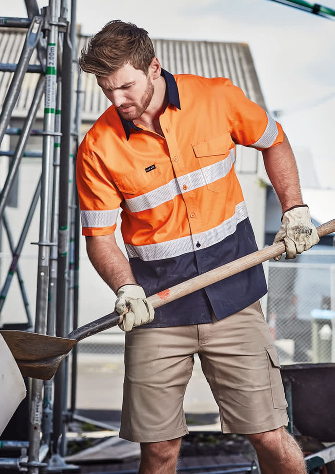 A short sleeve work shirt in orange and navy with reflective tape, worn by a man using a shovel, with a construction setting in the background.