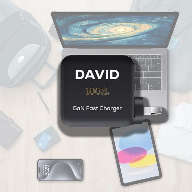 A 100W GaN quick charger in black, placed on a wooden desk alongside a laptop, tablet, and smartphone.