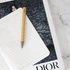 A bamboo pen with a metallic tip resting on a lined notebook, against a dark patterned background featuring a logo.