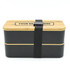 A two-layer lunch box with a dark exterior and a bamboo lid, secured with a black strap. Features a logo.