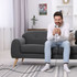 A man seated on a grey couch using a phone, with a charging cable visible on the floor. The setting is bright and modern.