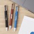 Three eco pens in black, red, and blue, with white tips, resting on a wooden surface alongside a notebook and laptop.