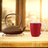 A red ceramic mug next to a decorative teapot on a wooden surface, set against a blurred background.