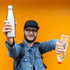 A drink bottle in stainless steel held by a person, against an orange background, with a plastic water bottle in the other hand.