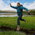 A drink bottle is held by a person in a blue jacket, jumping over a stream in a natural landscape.