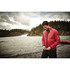 A red puffer jacket is worn by a man standing near a river and forested area, with a logo on the sleeve.