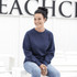 A woman sits wearing a navy blue fleece crew jumper, paired with light blue jeans, smiling against a neutral background.