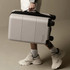 A grey carry-on suitcase with wheels being held by a person wearing white sneakers and ankle socks. The suitcase has a logo.