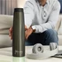 A drink bottle in a sleek, dark green colour with a logo, placed on a table beside headphones.