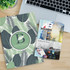 A medium notebook with a green leaf design alongside a brochure on a desk with a laptop and a plant.
