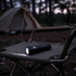 A black LED torch resting on a camping chair, with a tent in the blurred background among trees.