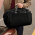 A black duffel bag with carrying handles is held by a person near a car trunk. The bag has a logo on it.