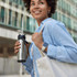 A drink bottle in stainless steel, held by a person wearing a blue shirt and carrying a tote bag. The bottle features a flip straw and has a logo.