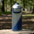 A drink bottle in grey and navy blue, featuring a large handle and a spout, placed on a rock in a natural setting.
